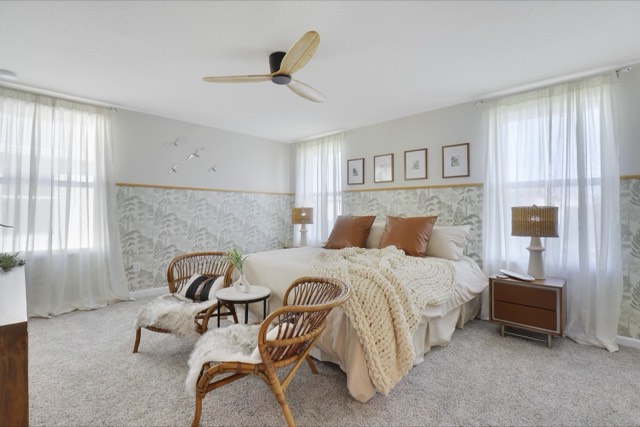 Primary bedroom with botanical wallpaper below a wood rail, sheer curtains on two windows and two rattan chairs