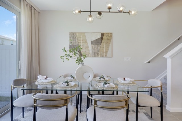 Glass dining table with cream chairs under a black linear chandelier, the sliding door at the left