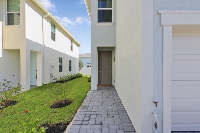 Paver walkway beside the garage leading to a dark front door, with lawn and shrubs at the left