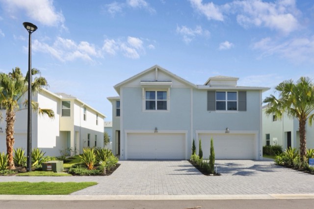 Front of the two-story blue-gray townhome building with two garage doors, palms and a paver driveway