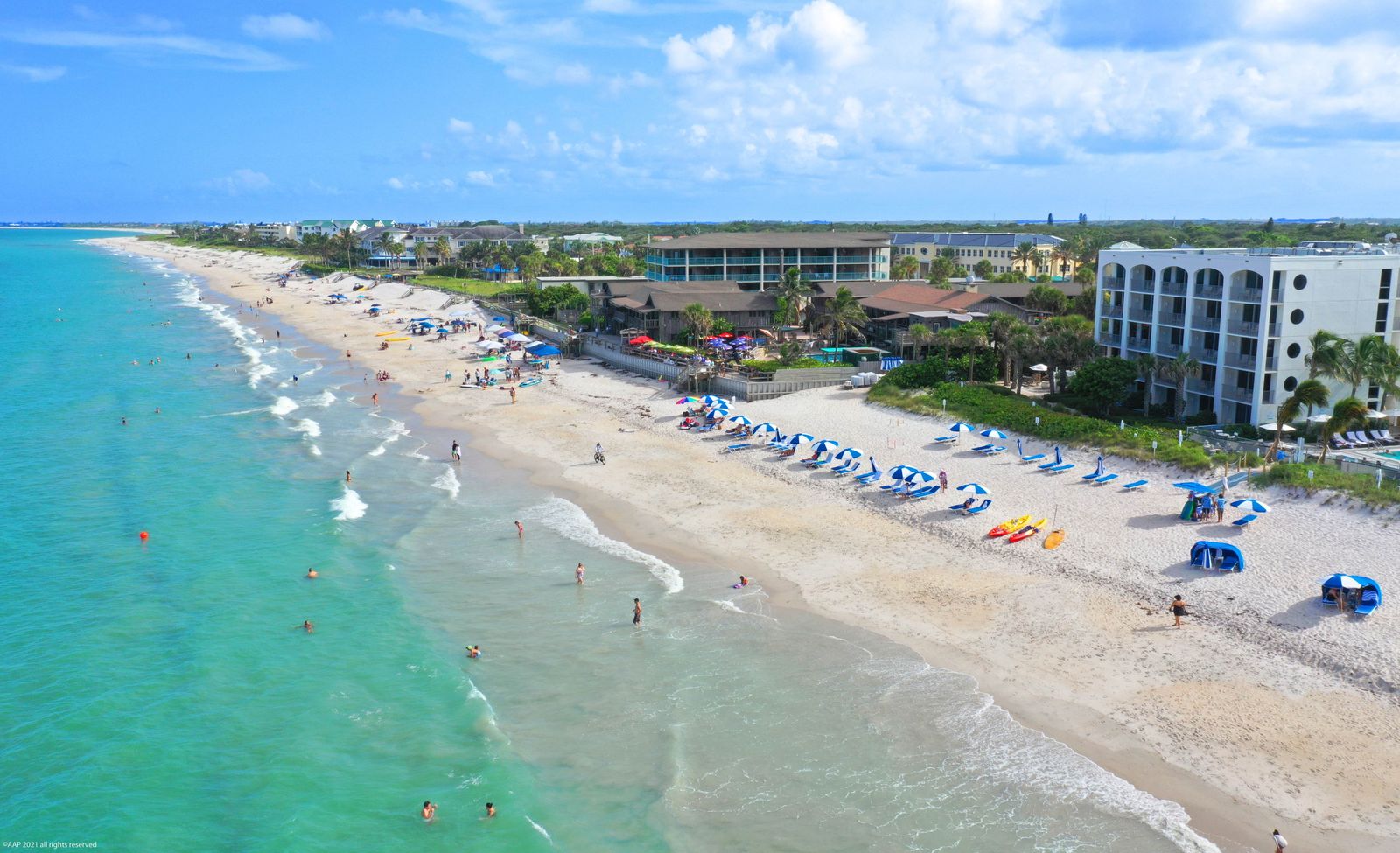Aerial of the Vero Beach shoreline, turquoise water, beachgoers and umbrellas, hotels behind the dunes